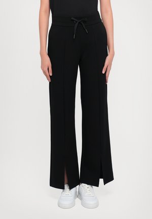 Personne portant un pantalon noir à jambes larges avec des fentes à l'avant et une taille à cordon, associé à des baskets blanches, debout devant un fond uni.