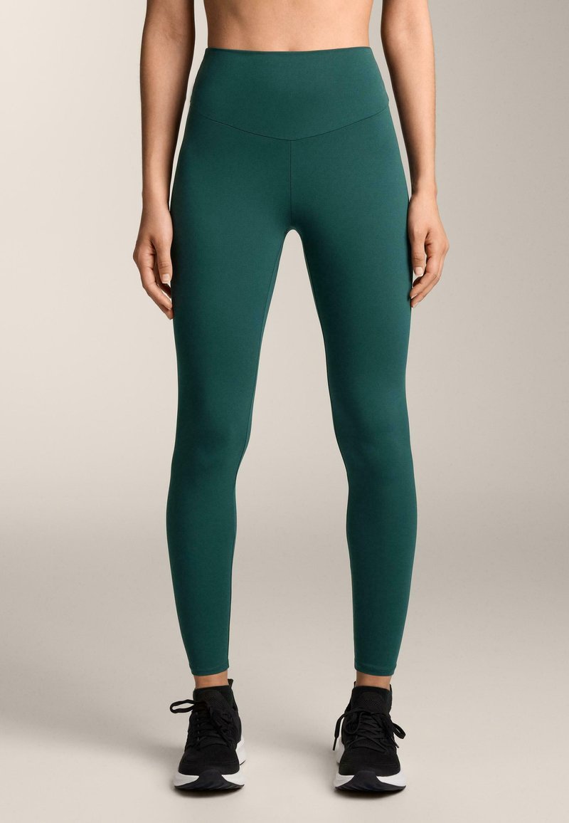Hoch taillierte, eng anliegende Leggings aus tiefgrünem Stoff, mit glatter Textur und minimalistischem Design. Dazu schwarze Sportschuhe kombiniert.