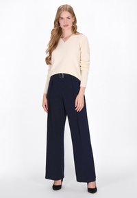 Pulover bej cu decolteu în formă de V, asortat cu pantaloni largi bleumarin, având o curea cu cataramă argintie, purtat cu tocuri negre, pe un fundal alb.