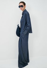 Completo in denim composto da una camicia con bottoni e taschino sul petto, abbinata a pantaloni a gamba larga. Gli accessori includono occhiali da sole grandi e una borsa scura.