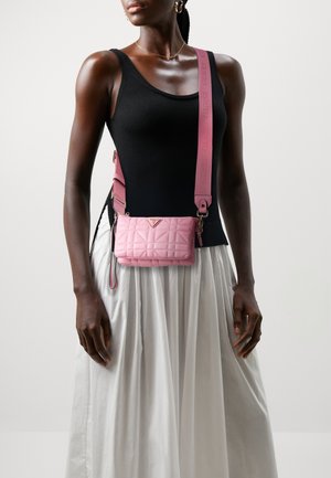 Różowa pikowana torebka na ramię z logo, z szerokim paskiem w tym samym kolorze. Nosi się ją na czarnym topie, zestawioną z białą spódnicą.