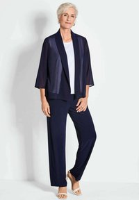 Marine doorzichtige kimono-jack over een witte top, gecombineerd met marineblauwe wijde broek. Geaccentueerd met sandalen, die een lichte textuur en een losse pasvorm tonen.