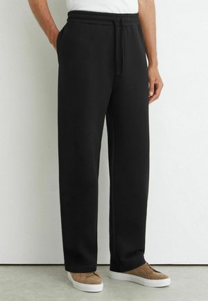 Person iført sorte sweatpants med snøre og brune sneakers, stående mod en hvid væg med hænderne i lommerne.