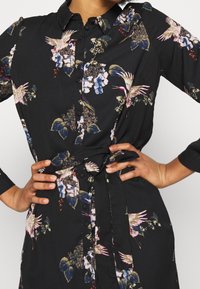 Abito a camicia floreale in tessuto nero, caratterizzato da motivi colorati di uccelli e fiori, maniche lunghe e una vita legata in evidenza.