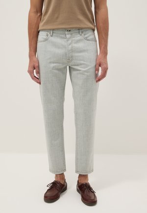 Hombre con vaqueros gris claro de corte recto, zapatos marrones de cuero con cordones y camiseta beige sobre un fondo liso.