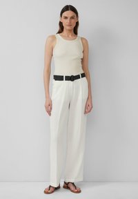 Beige ärmelloses Top kombiniert mit weißen, strukturierten weiten Hosen, akzentuiert durch einen schwarzen, gemusterten Gürtel und braunen flachen Sandalen.