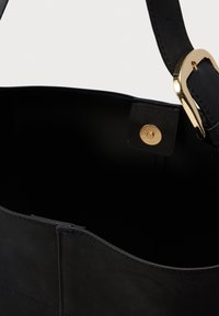 Indvendig på en sort lædertaske, der viser magnetlukning og en del af remmen med guldspænde på hvid baggrund.