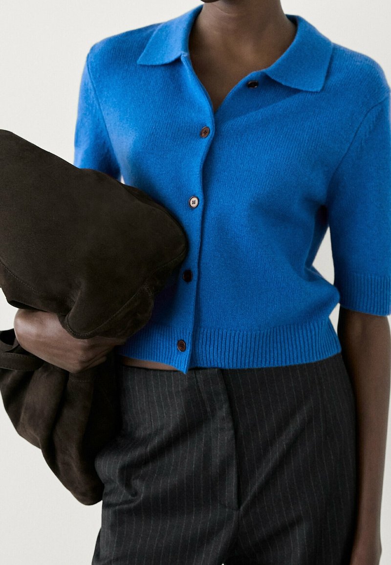 Personne portant un cardigan boutonné à manches courtes bleu vif et un pantalon rayé sombre, tenant une veste en suède brun foncé pliée.