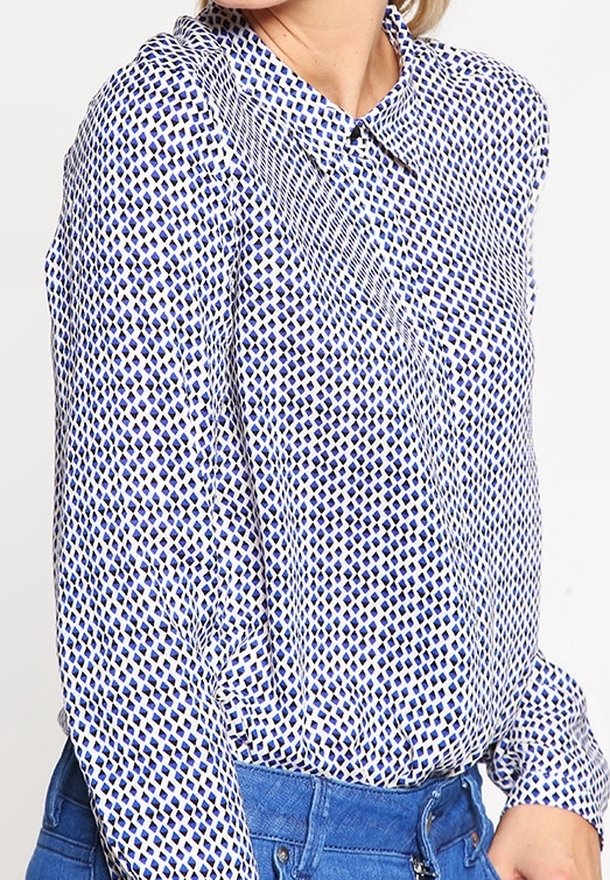 Chemise à manches longues blanche avec un motif géométrique bleu, dotée d'un col et d'une fermeture à boutons. Le tissu semble léger et lisse.