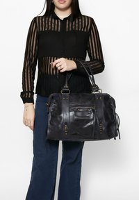 Borsa a tracolla in pelle nera con finitura texture, tasca con zip e accessori in metallo dorato; abbinata a una camicetta trasparente a righe nere e jeans blu.