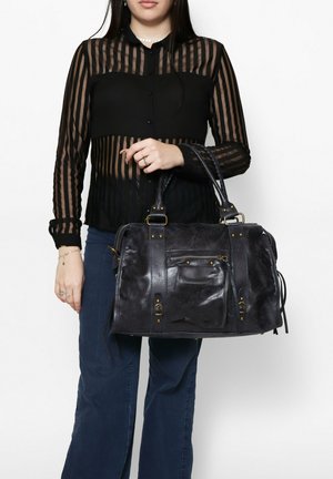 Borsa a tracolla in pelle nera con finitura texture, tasca con zip e accessori in metallo dorato; abbinata a una camicetta trasparente a righe nere e jeans blu.