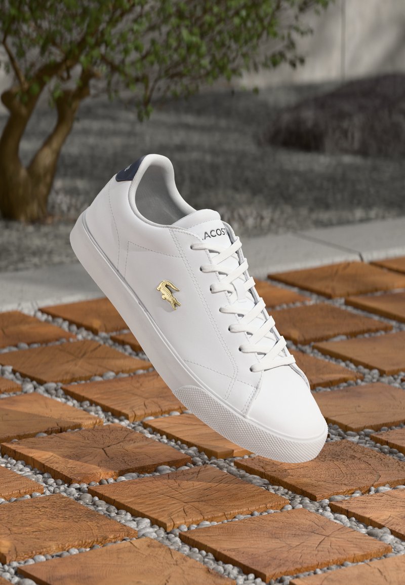 White low-top sneakers with smooth leather, a textured rubber sole, and gold Lacoste logo. Features lace-up front and navy heel accents.