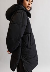 Schwarze Pufferjacke mit passgenauer Kapuze, horizontal gestepptem Design, seitlicher Reißverschlusstasche und einem etwas längeren Rückenbereich. Weiche, matte Textur.