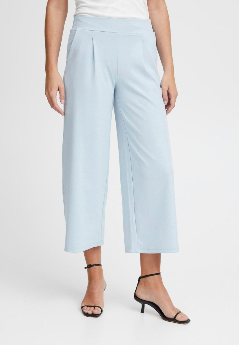 Lichtblauwe wijde broek met hoge taille en plooien, gemaakt van zachte stof. Gecombineerd met zwarte sandalen met bandjes voor een moderne uitstraling.