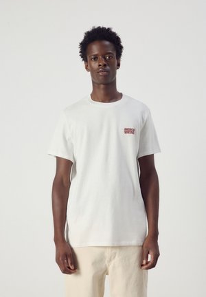 Jeune homme aux cheveux courts et bouclés portant un t-shirt blanc avec un petit texte rouge sur la poitrine et un pantalon beige clair, debout sur un fond uni.