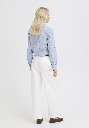 Pikade blondide juustega naine, kes kannab sinist triibulist särki väikeste lillemustritega, valgeid pükse ja pruune mokassiine, seisab valge tausta ees küljepealt.