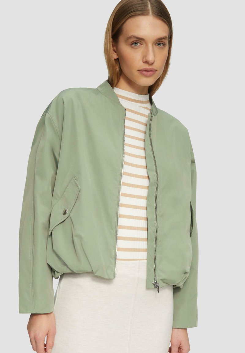 Giacca bomber verde chiaro con chiusura a zip, vestibilità comoda, tasche laterali e colletto alto, indossata sopra una maglietta a righe. Tessuto dalla texture liscia.