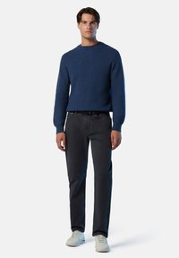 Suéter de punto texturizado en azul con cuello redondo, combinado con pantalones gris oscuro y zapatillas gris claro, de pie frente a un fondo blanco.