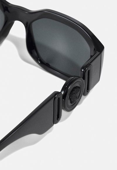Black sunglasses with a chunky frame, dark lenses, textured temples, and an embossed emblem on the side. Smooth finish with modern design elements.