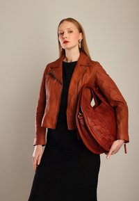 Kvinde iført en brun læderjakke og sort kjole holder en stor brun vævet lædertaske mod en beige baggrund.