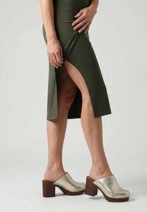Vrouw die metalen zilveren houten klompen draagt en een olijfgroene midi-rok met dijhoge split, staand tegen een effen achtergrond.