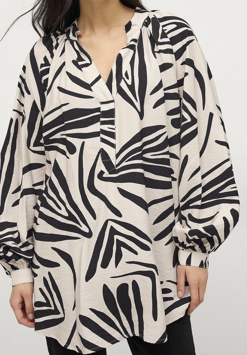Lettet bluse i cremefarvet stof med markante sorte zigzag-mønstre, V-hals og lange puffærmer med tætsiddende manchetter.