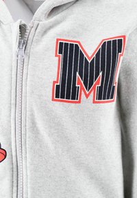 Grauer Zip-Hoodie mit einem großen schwarzen und roten gestreiften "M"-Logo. Glatte Textur, sichtbares Reißverschlussdetail an der Vorderseite.