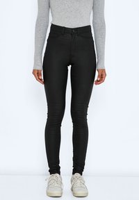 Personne portant un pantalon skinny noir taille haute, un haut gris à manches longues et des baskets blanches, debout devant un fond uni.