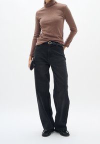 Femme portant un pull à col roulé marron, une ceinture noire, un jean noir ample, des chaussures noires, tenant une pochette noire sur un fond uni.