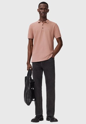 Mand iført en lysebrun poloshirt og sort jeans, der holder en sort læderrygsæk, stående mod en ensfarvet lys baggrund.