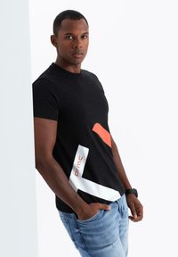 Musta t-paita, jossa on oranssi ja valkoinen abstrakti graafinen muotoilu. Lyhyet hihat ja pyöreä kaula-aukko. Yhdistetty vaaleansinisistä farkuista.