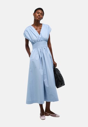 Femme portant une robe midi bleu clair sans manches avec une taille froncée, tenant un sac à main noir froncé et portant des chaussures plates rose clair.