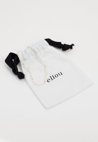 Sacchetto di tessuto bianco con lacci neri, con il testo "éliou." Una delicata collana di piccole perle rotonde è esposta sopra.