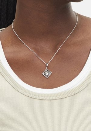 Collier en argent avec un pendentif carré présentant un cœur découpé et des maillons de chaîne, porté par une personne portant un haut côtelé beige avec un col blanc.