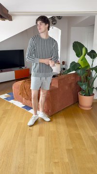 Ριγέ μακρυμάνικη μπλούζα σε μαύρο και λευκό, συνδυασμένη με γκρι σορτς. Λευκά αθλητικά. Εσωτερικός χώρος με φυτό και καναπέ.