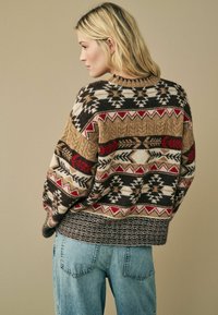 Pull en tricot avec des motifs géométriques dans des tons de marron, rouge, beige et blanc, doté un large ourlet côtelé. Associé à un jean en denim bleu clair.