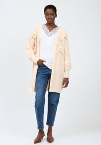 Strik cardigan i blød ferskenfarve med blomsterstruktur og åben front. Båret over en hvid top med blonder, parret med blå jeans og brune mules.