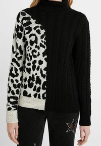 Pull à col roulé noir et crème avec un design en tricot torsadé et imprimé léopard, doté de poignets et d'un ourlet côtelés. Style texturé et asymétrique.