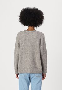 Gai pelēks, masīvs adījuma džemperis ar brīvu piegriezumu, ar ribotām piedurknēm un noapaļotu kakla izgriezumu, kopā ar gaiši ziliem džinsiem.