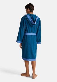 Personne portant un peignoir à capuche bleu, arrivant au genou, avec des bordures et une ceinture bleu clair, se tenant pieds nus face à un fond blanc.