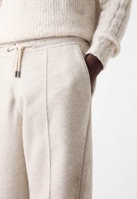 Beige jogger kalhoty z vlněné směsi mají boční kapsy a stahovací pas s kovovými detaily. Měkká textura a přizpůsobený střih.
