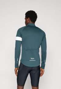 Chaqueta de ciclismo para hombre en color azul verdoso con mangas largas, banda de acento blanca en la manga, bolsillos traseros y marca en el dobladillo. Lleva puesta sobre pantalones cortos de ciclismo negros.