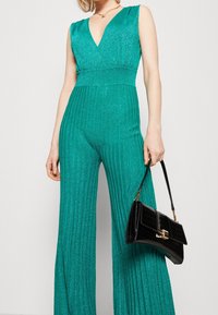 Mono de purpurina turquesa con escote en V, textura plisada y piernas anchas. Combinado con un bolso negro de piel de cocodrilo.