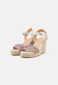 Sandales wedge argentées avec des brides multicolores tressées, talons en jute et semelles en cuir tan. Équipées d'une sangle ajustable à la cheville.