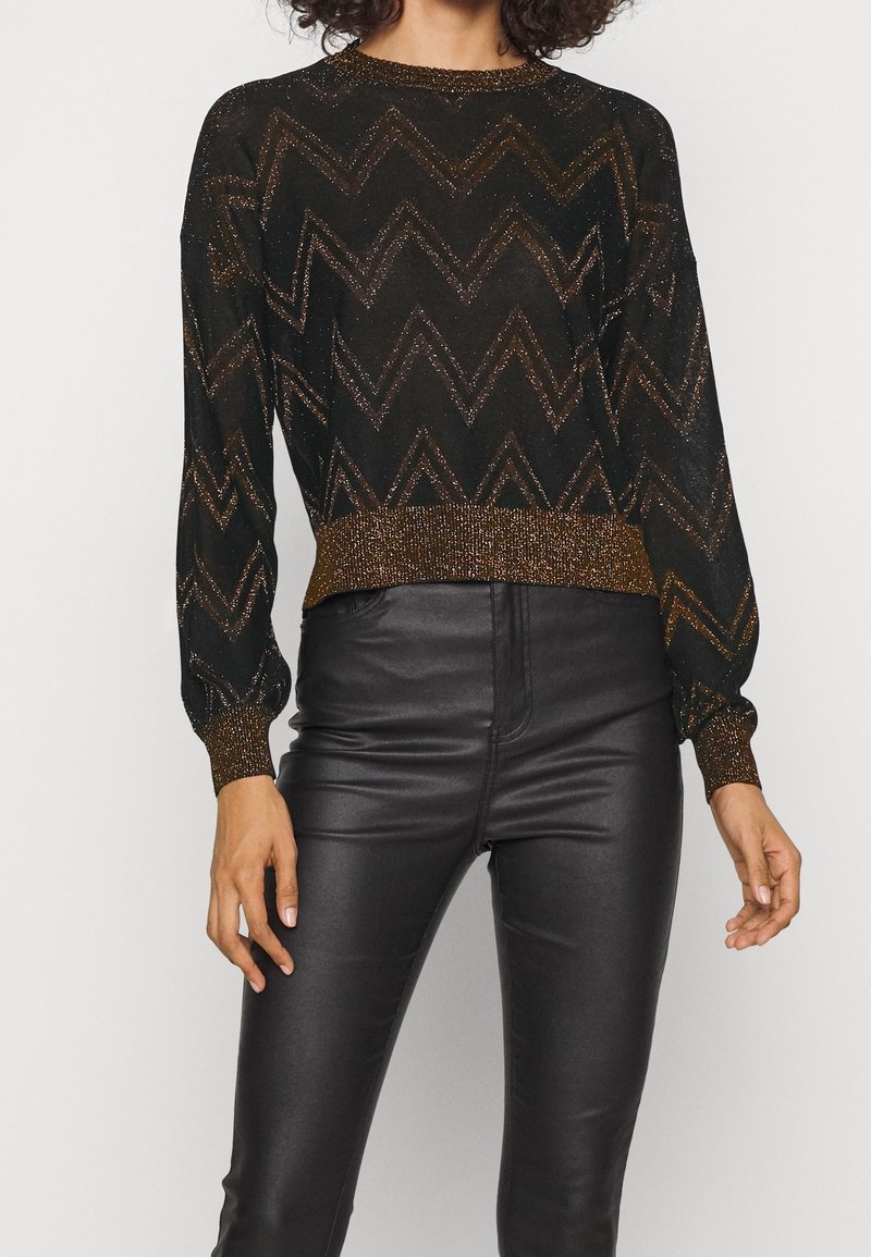 Femme portant un pull noir avec des motifs en zigzag scintillants et des poignets bronze, associé à un pantalon en simili-cuir noir sur fond blanc.