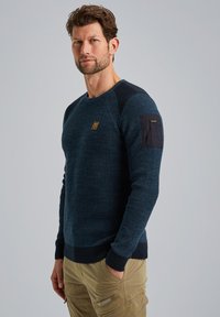 Marineblauer gestrickter Sweatshirt mit strukturiertem Stoff, gerippten Bündchen und Saum, versehen mit einem kleinen Logo-Patch am oberen linken Arm.