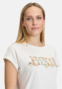 T-shirt en coton blanc à manches retroussées, ornée d'une broderie colorée "FIESTA" au centre. Texture douce et coupe décontractée.