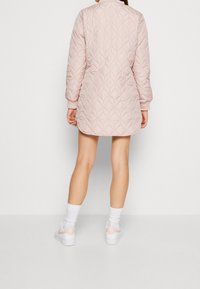 Polstret quiltet jakke i blød pink med diamantmønster, med rund krave og ribbede manchetter, båret over shorts og hvide sokker.