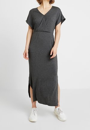 Vrouw draagt een antracietgrijze, korte mouwen, enkellange jurk met zijsplitten en witte sneakers, staand tegen een effen lichte achtergrond.