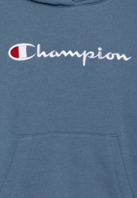 Sudadera con capucha azul hecha de mezcla de algodón, con un bolsillo frontal y el logo "Champion" bordado en blanco con un detalle en rojo.